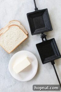 Slices of bread being buttered, next to a cast iron pie maker and a stick of butter
