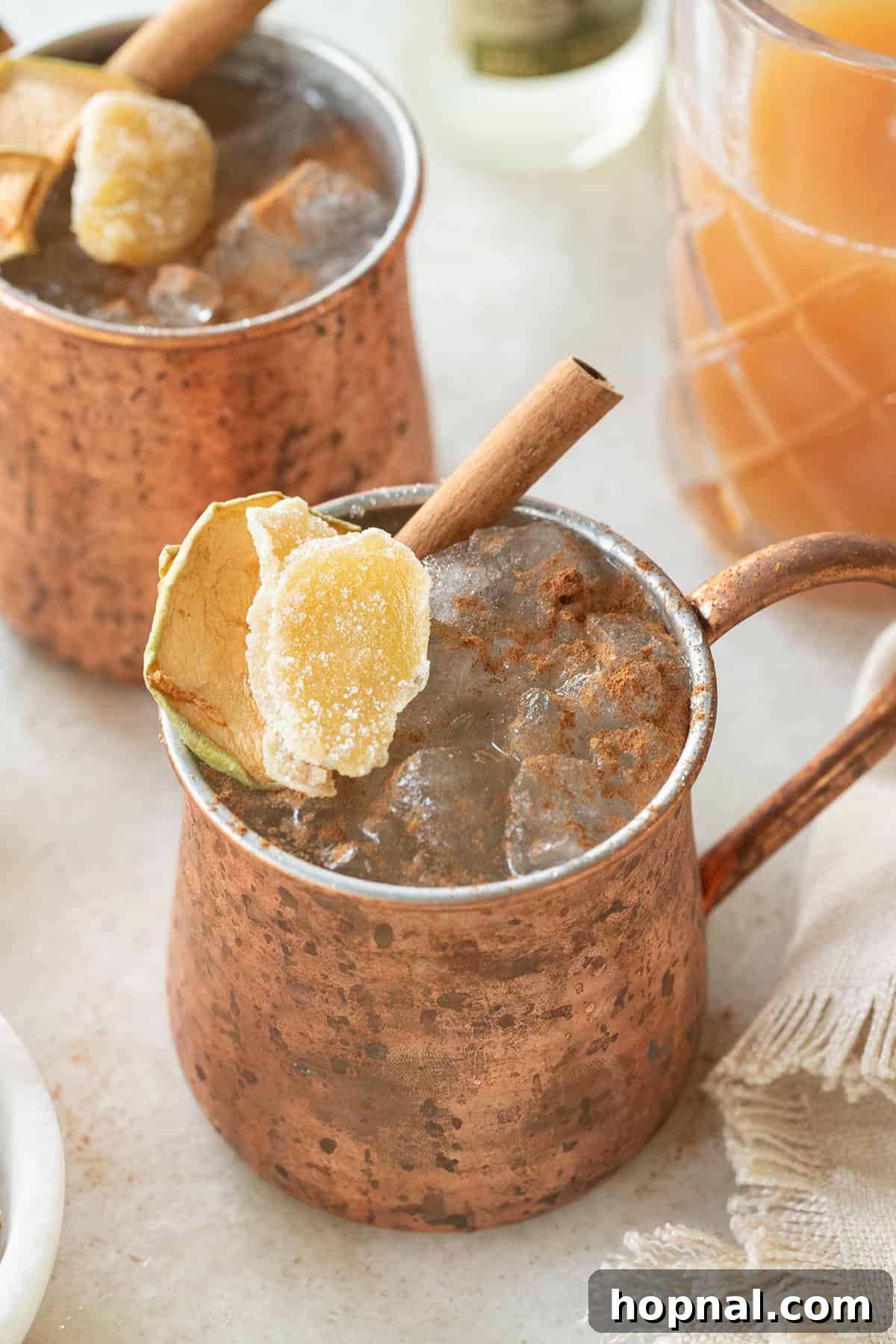 Spiced Apple Cider Moscow Mule 2 Icy Apple Cider Moscow Mule with a cinnamon stick and fresh apple slice garnish, served in a classic copper mug on a rustic wooden table with scattered autumn leaves.