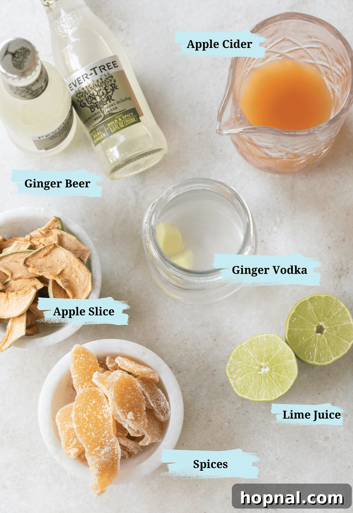 Spiced Apple Cider Moscow Mule 3 A vibrant flat lay of Apple Cider Moscow Mule ingredients: ginger beer bottles, a pitcher of apple cider, a bottle of ginger vodka, fresh lime halves, and a bowl of candied ginger for garnish.