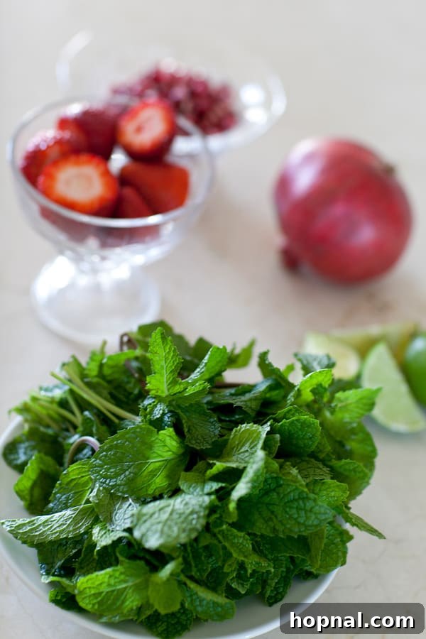A bowl of fresh mint leaves, with strawberries and a pomegranate in the background, essential for mojito preparation.
