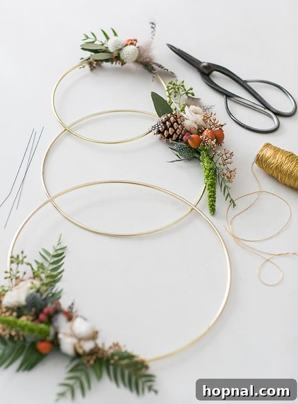 A stunning modern Christmas wreath composed of three interconnected gold metal rings, tastefully decorated with sparse green foliage and small pinecones, ready to hang.