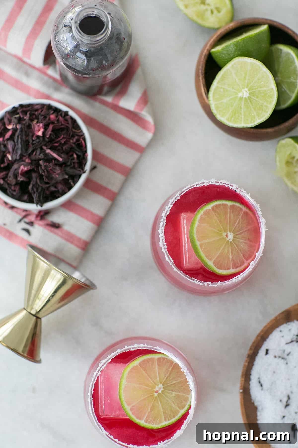 A close-up shot of a vibrant pink hibiscus margarita on a table, surrounded by fresh limes and dried hibiscus flowers, highlighting its natural beauty and ingredients.