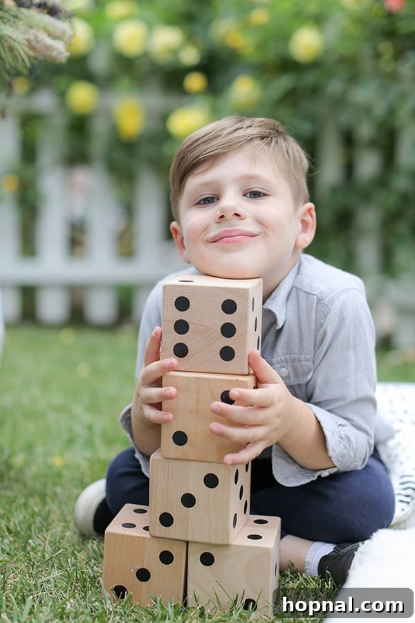 A cheerful little boy holding large wooden dice, ready for a game at a lively outdoor picnic gathering.