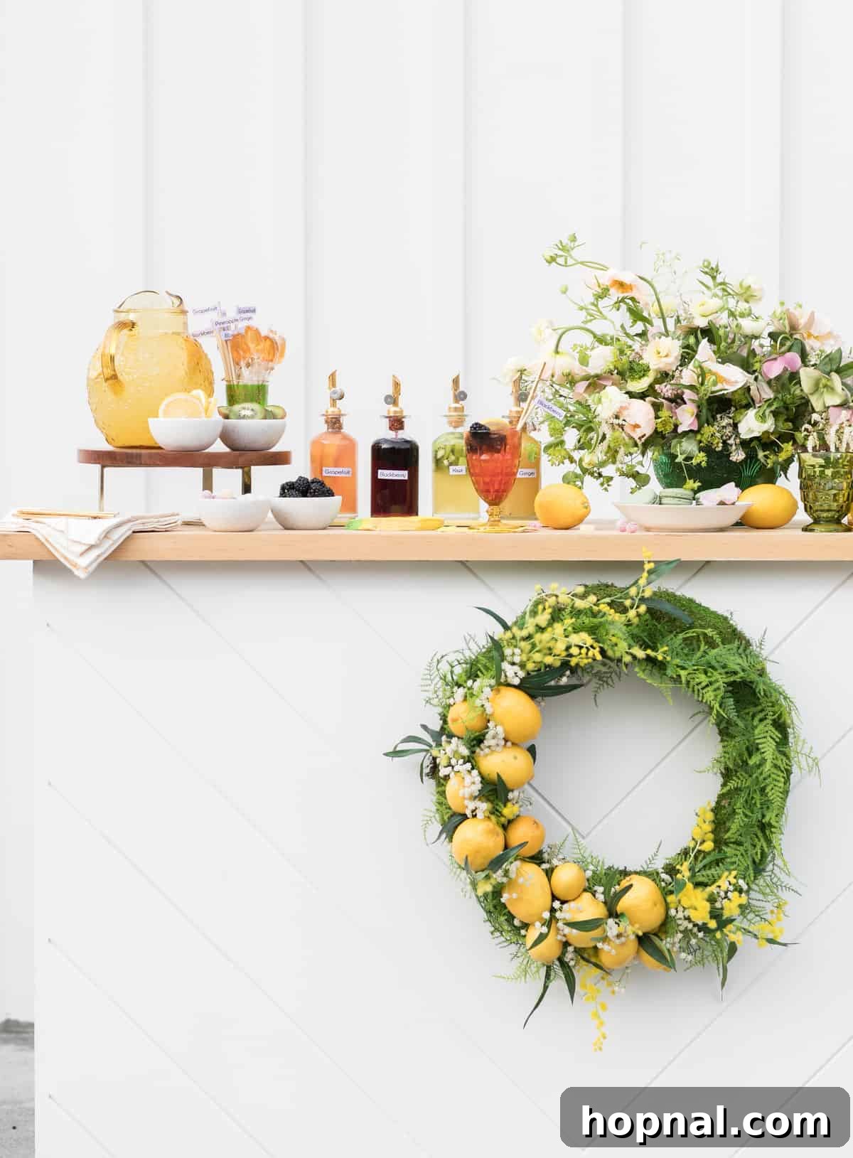Elegant white lemonade stand adorned with fresh lemons, a festive wreath, and an assortment of homemade fruit syrups, ready for an adult summer party.