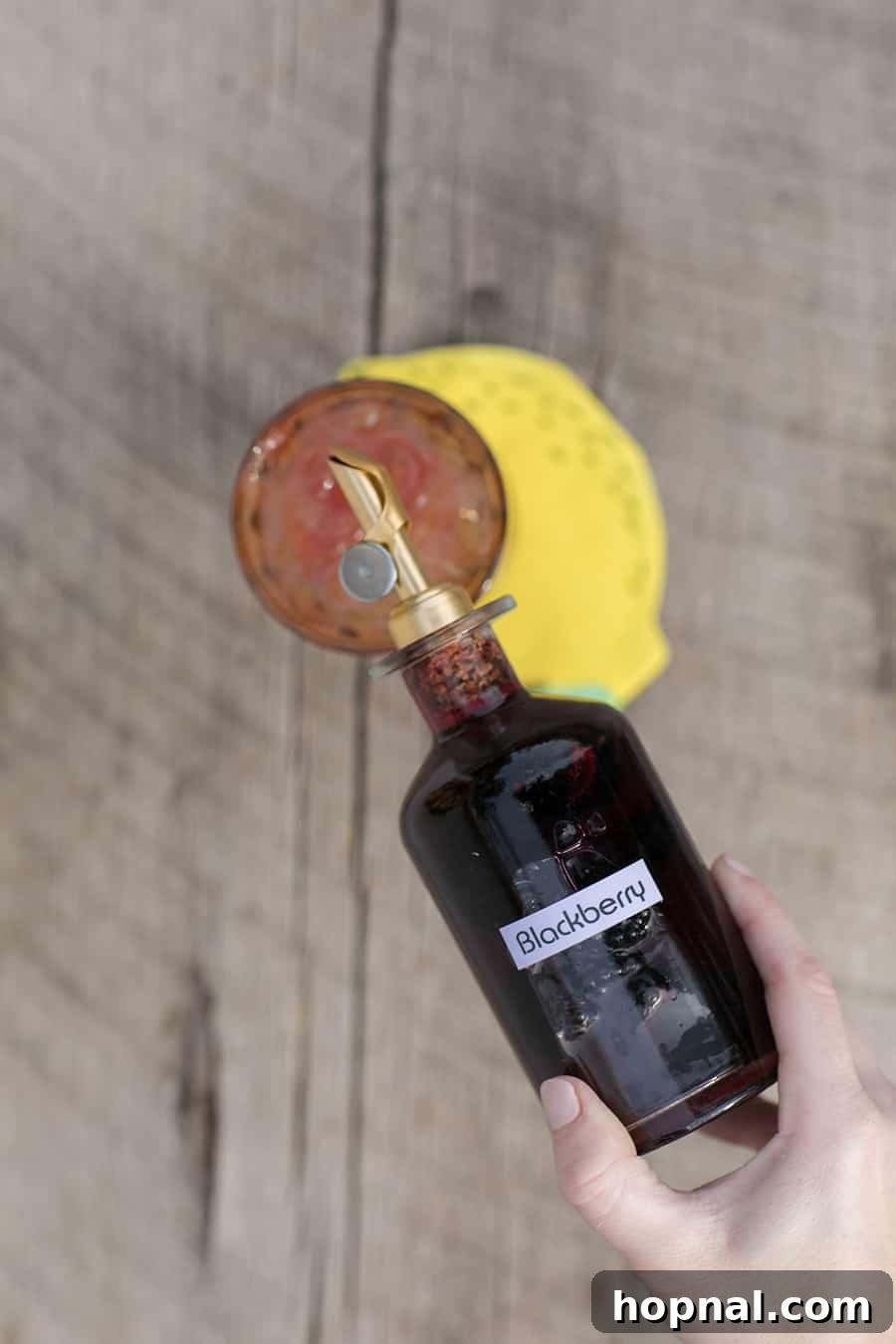 A hand pouring rich, dark blackberry syrup from a stylish bottle with a gold spout into a glass, illustrating the customization process of making a spiked lemonade.