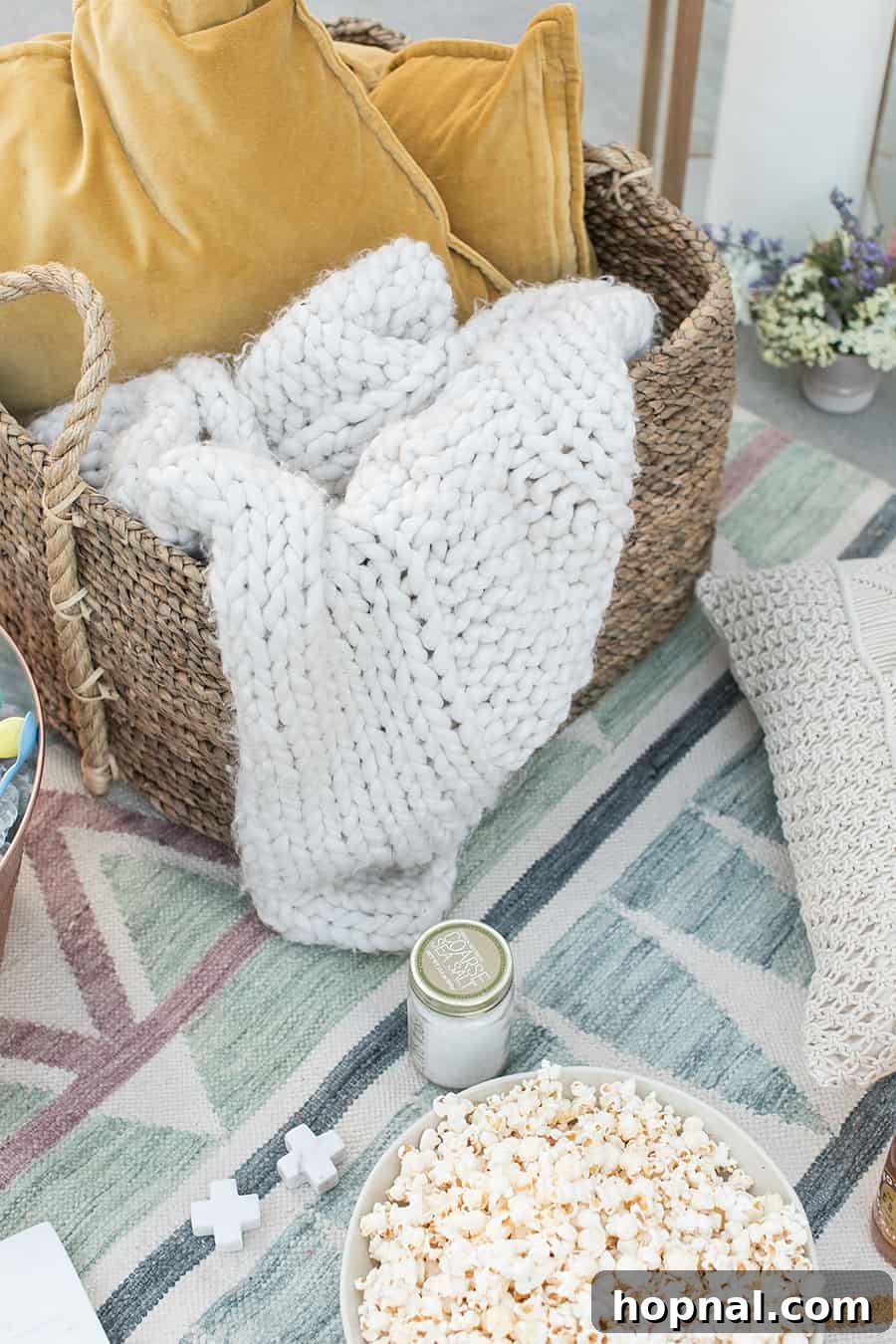 Lights, Camera, Backyard: Your Outdoor Movie Night Guide 5 A woven basket filled with a soft knitted blanket, alongside a bowl of popcorn, resting on a modern blue outdoor rug.