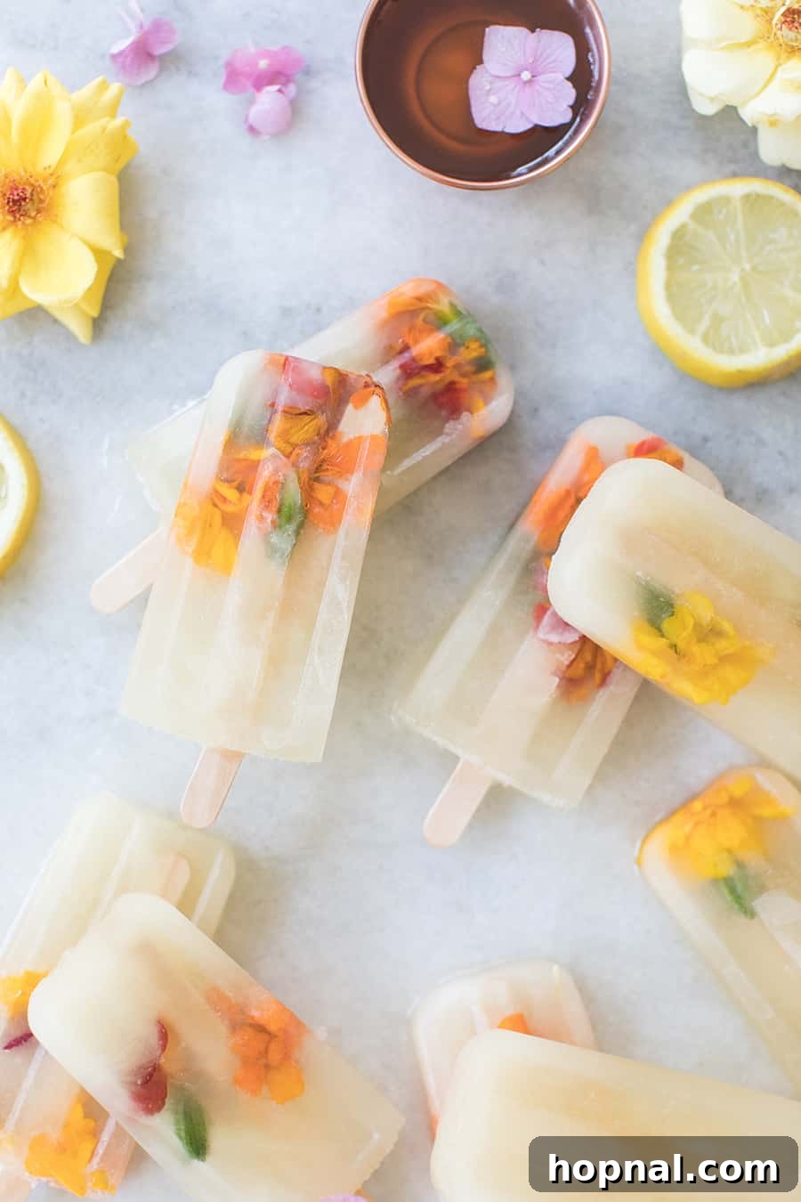 Three Ingredient Honey Lemonade Popsicles artfully arranged with edible flowers and fresh lemons on a pristine marble table.