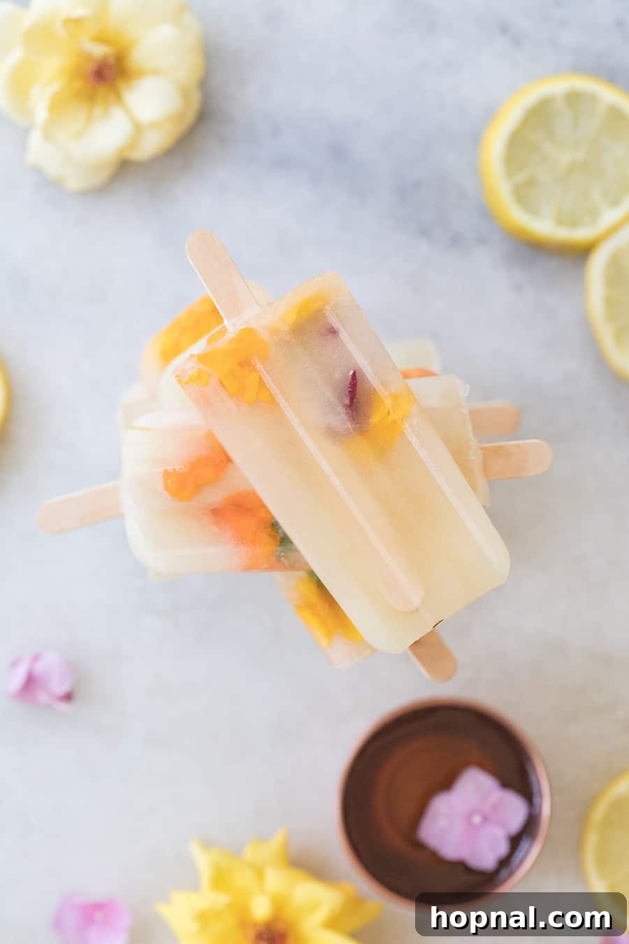 Three Ingredient Honey Lemonade Popsicles with edible flowers and lemons on a marble table, showcasing the finished product.
