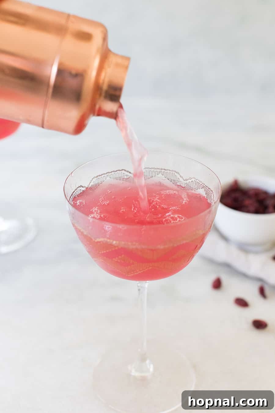 Pouring a pink cranberry cocktail into a coupe glass, showcasing its vibrant color and elegant presentation.