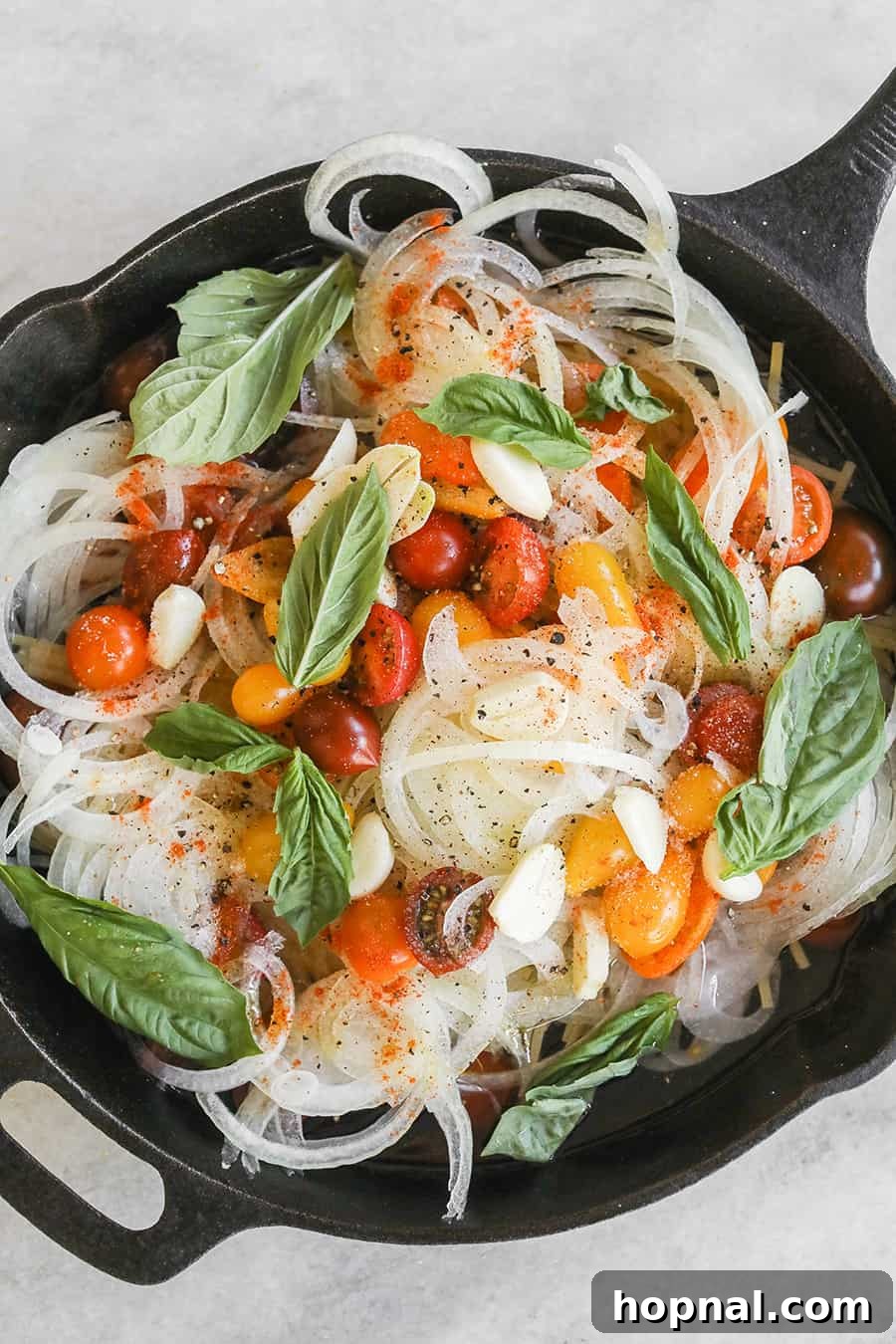 Dry linguine pasta, sliced garlic, halved cherry tomatoes, and thinly sliced onions arranged in a cast iron pan, ready for cooking the one pan basil and tomato pasta.