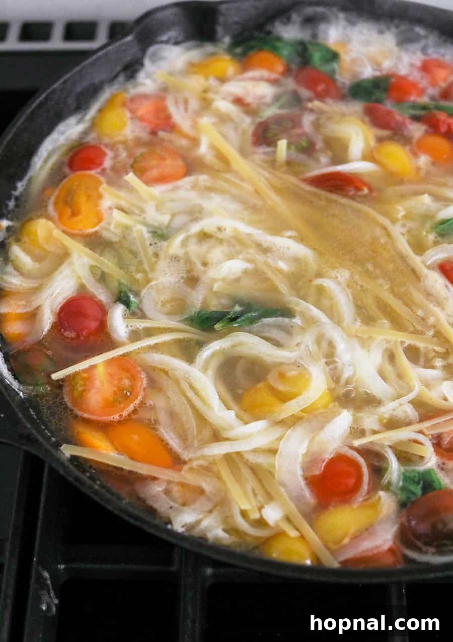 One Pan Basil and Tomato Pasta actively cooking in a cast iron pan, showing the pasta simmering with tomatoes, basil, and onions in a flavorful liquid.