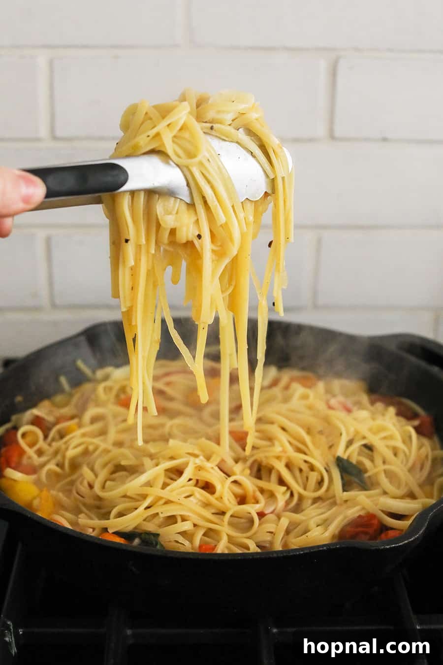 Close-up of tongs lifting a handful of cooked linguine pasta with tomatoes and basil above a cast iron skillet, showcasing the perfect texture and sauciness of the one pan dish.