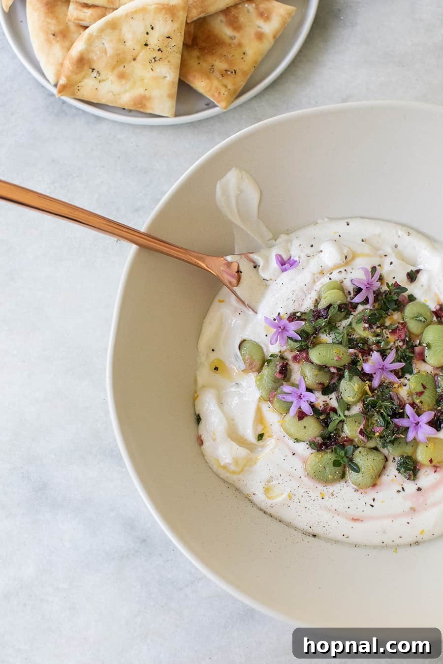 A white bowl brimming with creamy yogurt tahini dip, artfully topped with colorful lima beans, fresh thyme, and a drizzle of olive oil, ideal for sharing.