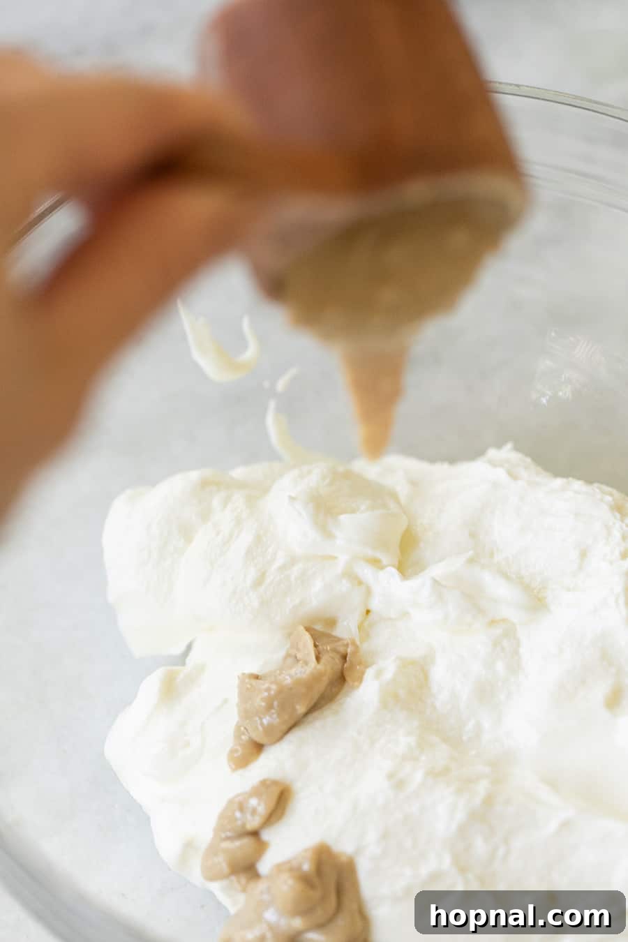 Smooth tahini paste being poured from a measuring cup into a bowl of creamy strained yogurt, illustrating the blending process for the dip base.