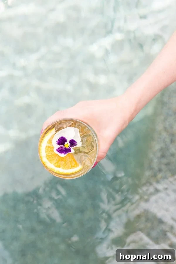 Hand holding a glass of pineapple party punch over a pool. It's garnished with an orange rind and edible flower.