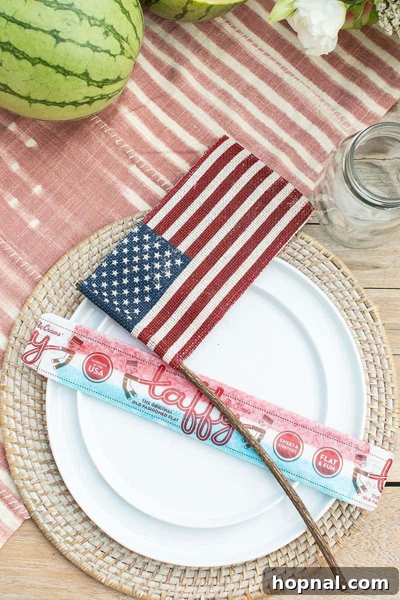 A festive 4th of July table setting with white plates on woven chargers, burlap American flags, and old-fashioned taffy candies, exuding a rustic yet elegant patriotic charm.