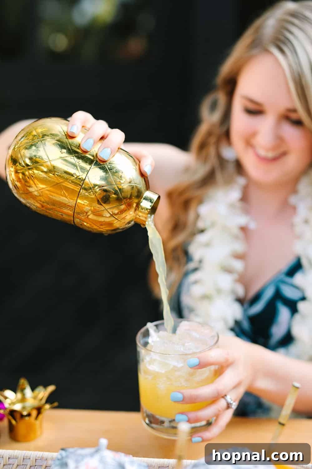 A person pouring a vibrant tropical cocktail into a glass, using a stylish gold pineapple cocktail shaker, highlighting drink preparation.