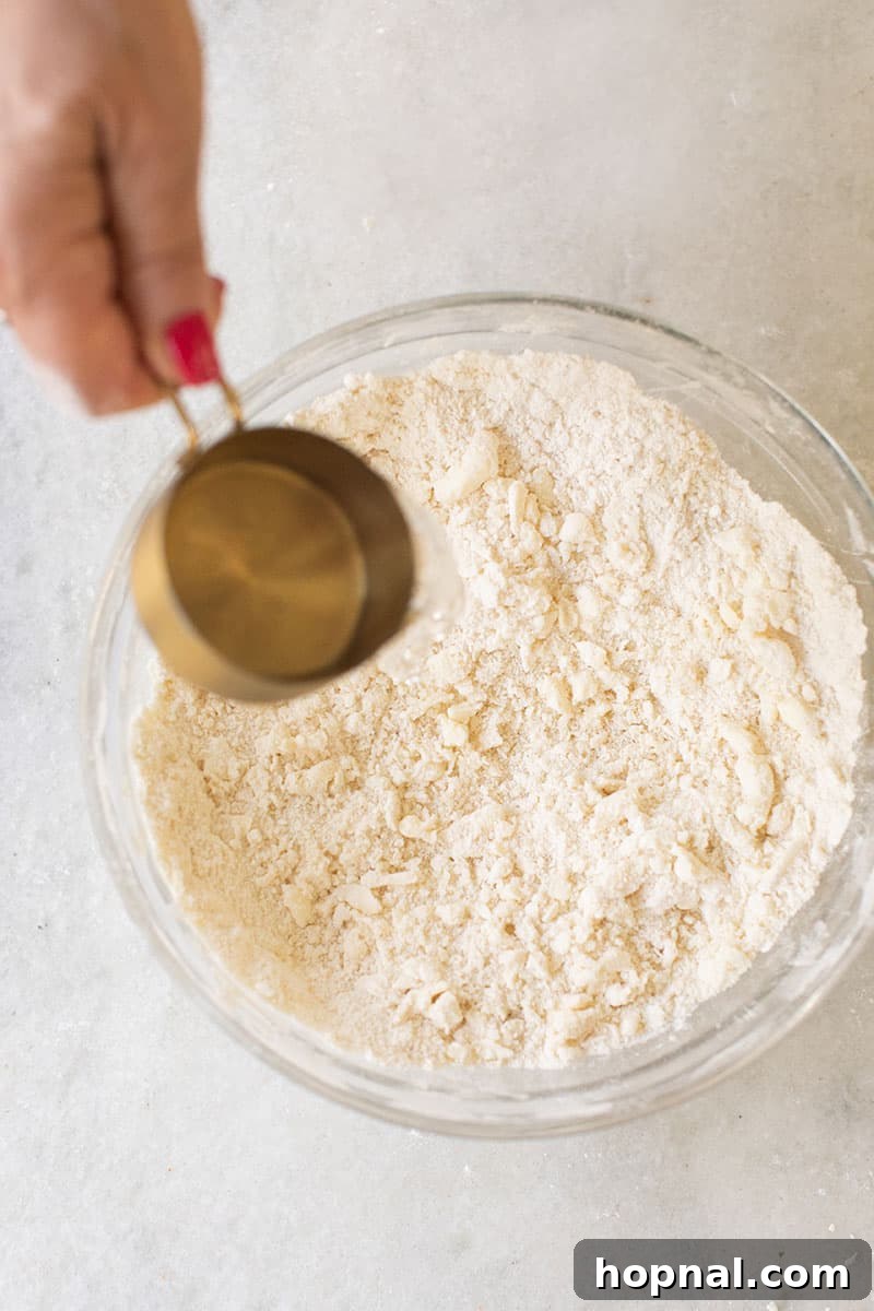 A hand carefully pouring ice water from a glass into a bowl of pie crust mixture, emphasizing the critical 'cold ingredients' rule.