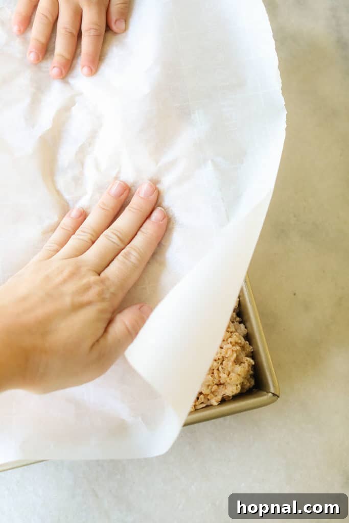 Using a piece of parchment paper to firmly press the Rice Krispie mixture into the pan for an even layer.
