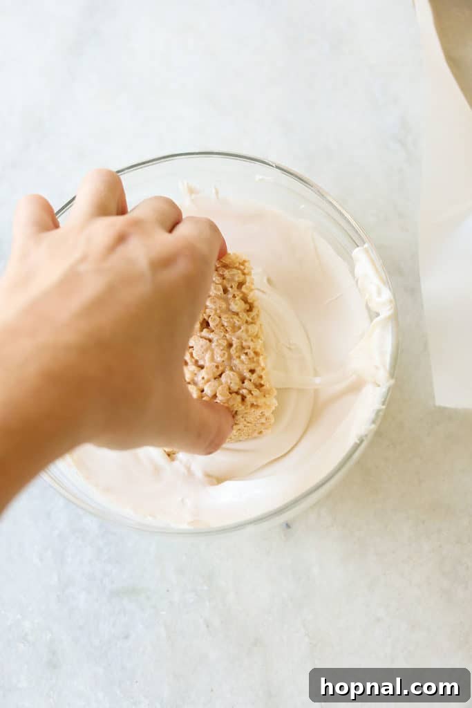 Dipping Rice Krispie treats into a bowl of melted white chocolate to coat them.
