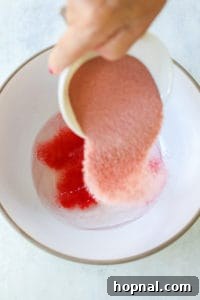 Adding red jello powder to a bowl with vodka and gelatin mixture.