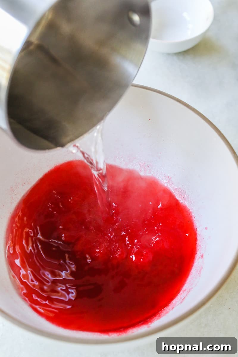 Boiling water is carefully poured into a white bowl containing red jello, vodka, and gelatin, all being continuously whisked to combine.