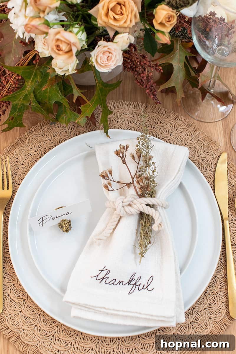 A close-up of a Thanksgiving table setting, featuring a linen napkin embroidered with 'thankful,' secured with a rustic rope and elegant gold silverware.