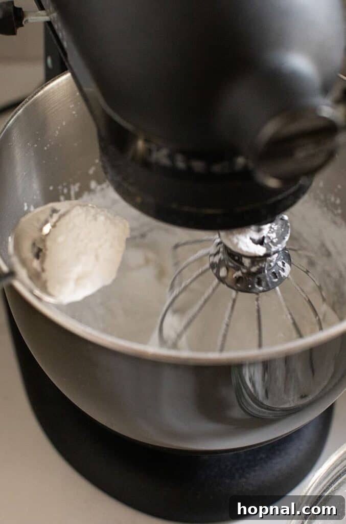 Powdered sugar being carefully added into a stand mixer bowl containing whipped egg whites.