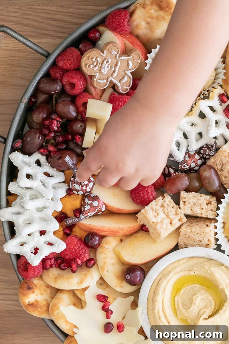 Santa's Snack Tray for Kids 3 A small child's hand gently reaching to pick up a classic Hershey's Kiss from a bountiful kids' Christmas appetizer platter, showcasing easy access and tempting treats for young party-goers.