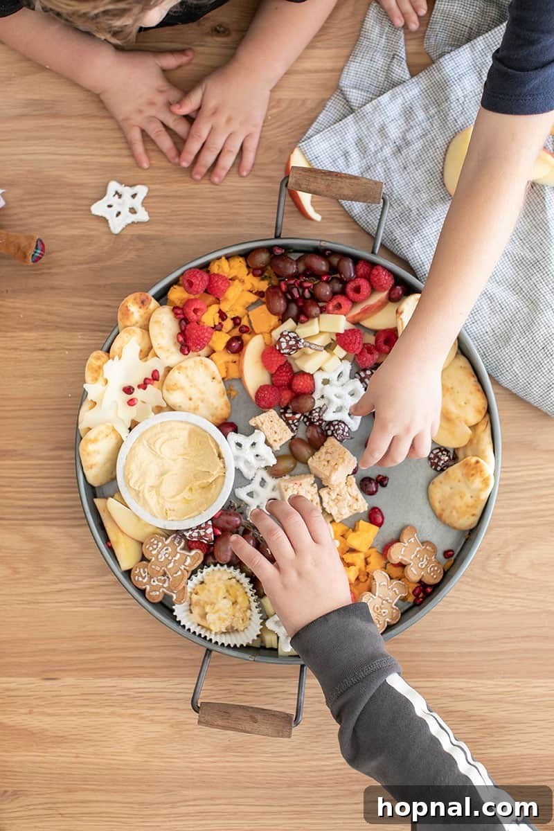 Santa's Snack Tray for Kids 9 Children eagerly reaching to select their favorite foods from a round galvanized tray, abundantly filled with a delicious assortment of creamy hummus, savory cheese, soft pita bread, crisp apple slices, sweet yogurt-covered pretzels, and classic gingerbread cookies, perfect for a kids' holiday party.