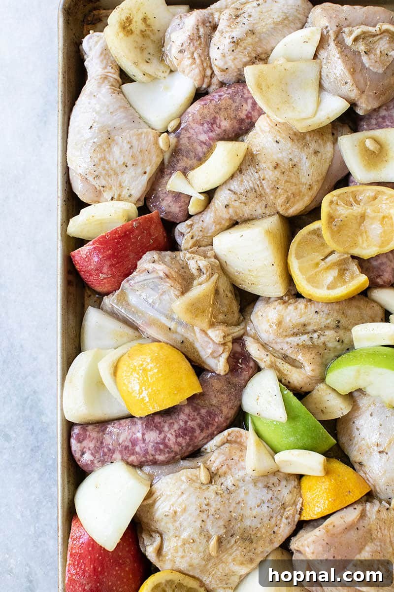 Raw chicken, Italian sausage, apples, lemons, and onions spread on a gold sheet pan, pre-bake.