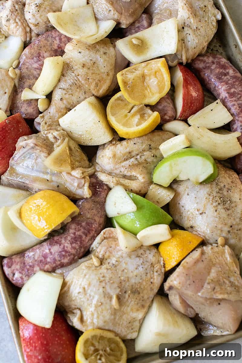 Garlic cloves, chicken, lemons, apples, and sausages beautifully arranged on a sheet pan before baking.