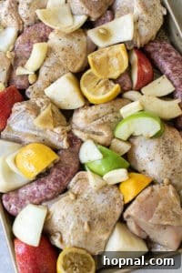 Raw chicken, sausage, apples, lemons, and onions arranged on a gold sheet pan before baking.