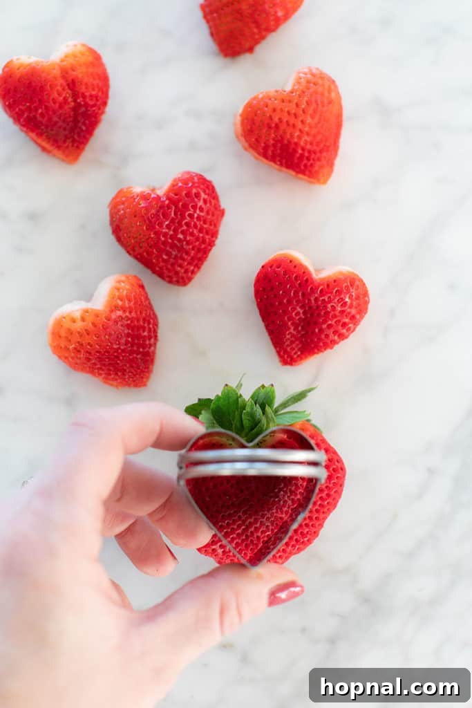 Valentine's Day Sweetheart's Grazing Board 17 A hand holding a heart-shaped cookie cutter, preparing to cut shapes from a large, ripe strawberry for a festive food decoration.