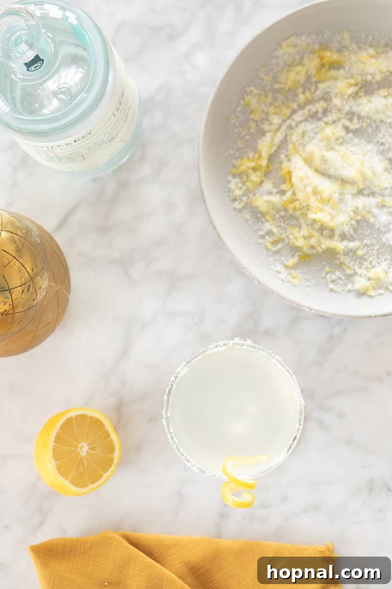 A beautifully prepared Lemon Drop Martini, accompanied by a lemon-sugar rim, rests on a marble table alongside a cocktail shaker and a bowl of fresh lemon sugar.