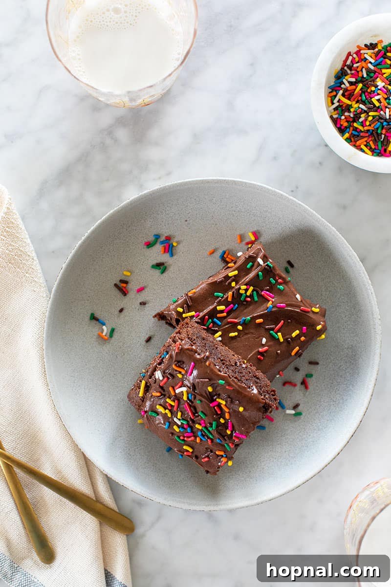 A delicious homemade brownie, beautifully frosted and adorned with vibrant rainbow sprinkles, resting on a crisp white plate, ready for a festive treat.