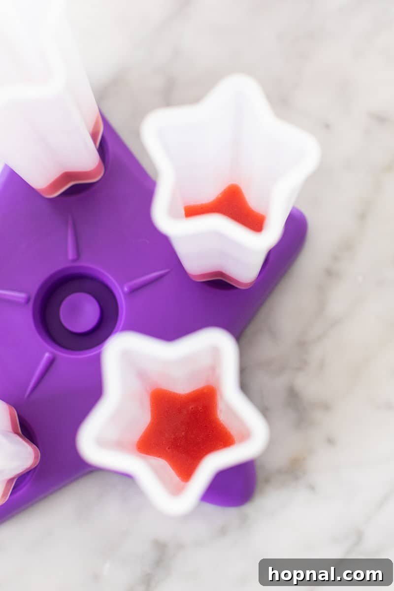 Vibrant red strawberry popsicle mix being carefully poured into a clear popsicle mold, forming the first layer.