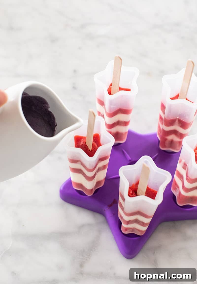 The final purple blueberry popsicle mixture being carefully poured into a mold already containing red and white layers, completing a striped popsicle.