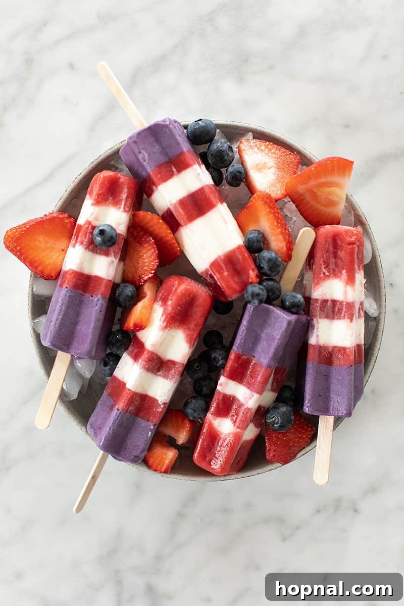 Homemade strawberry and blueberry layered popsicles arranged on ice, garnished with fresh strawberries and blueberries.
