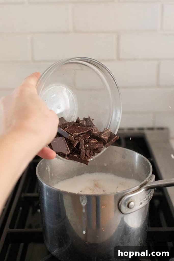 Pouring finely chopped dark chocolate into a saucepan containing warm heavy cream, preparing to melt into a smooth mixture.