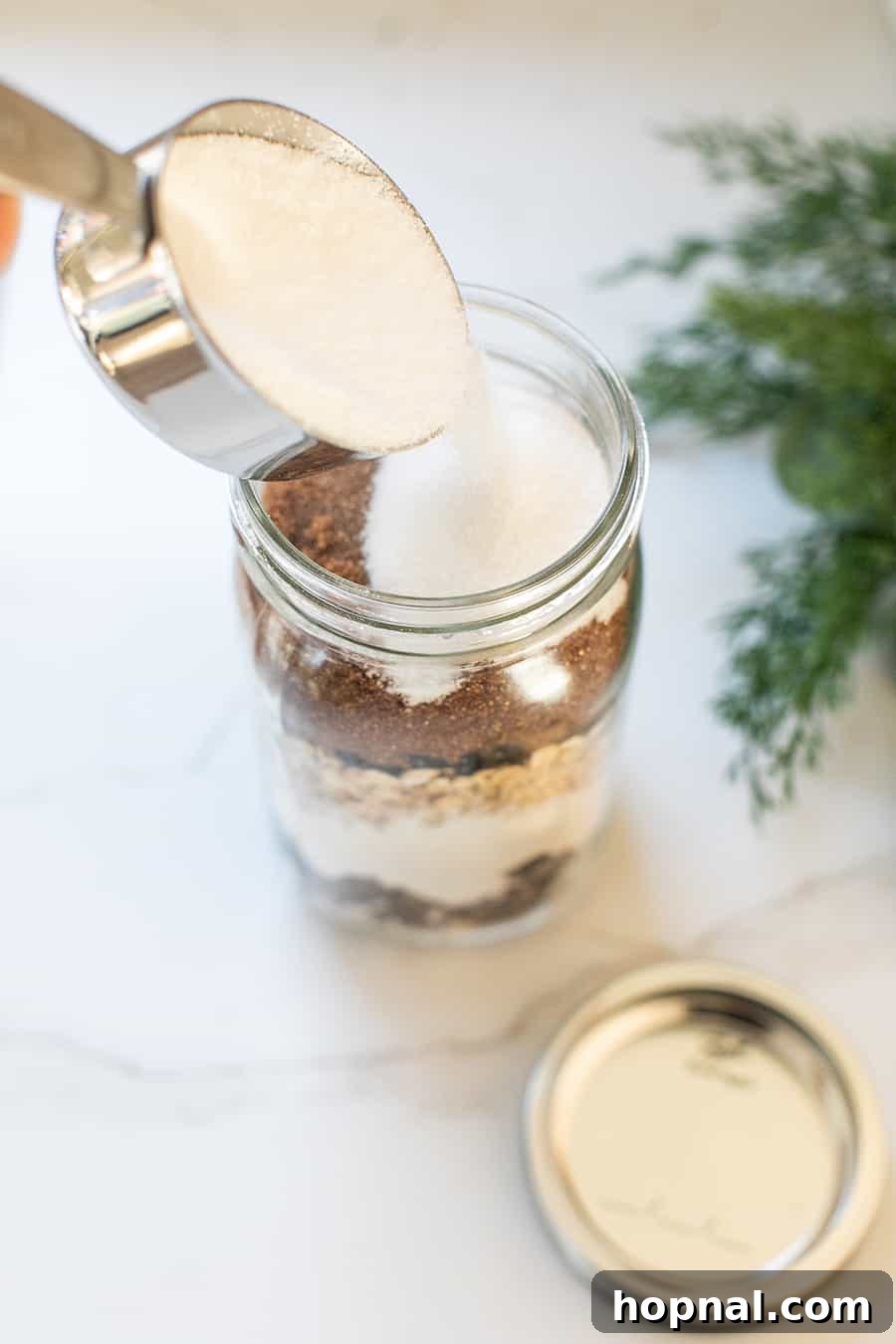 Granulated sugar being carefully poured as the top layer of a cookie mix in a jar, completing the Chocolate Cherry Chunk recipe.