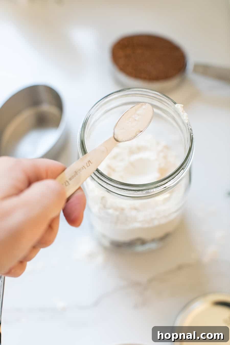 Dry ingredients, including flour and baking powder, being carefully added as a layer to a glass jar for the butter-less chocolate chip cookie mix.