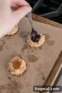 A close-up of partially baked thumbprint cookies on a baking sheet, with a small spoon carefully adding sweet jam into the indentations before the final bake.