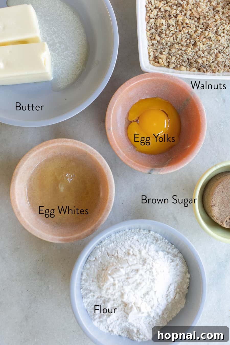 Bowls of essential ingredients for jam thumbprint cookies: butter, separated egg yolks and whites, all-purpose flour, finely crushed walnuts, and dark brown sugar, arranged on a classic marble surface.