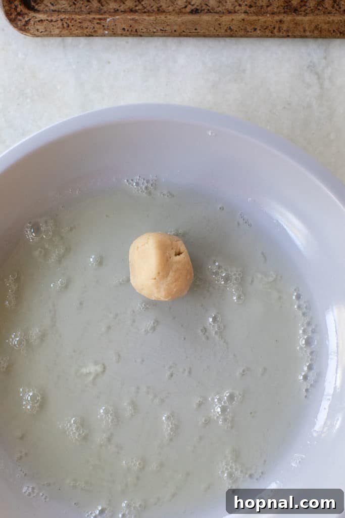 Cookie dough balls being rolled in egg whites in a bowl, preparing for walnut coating.