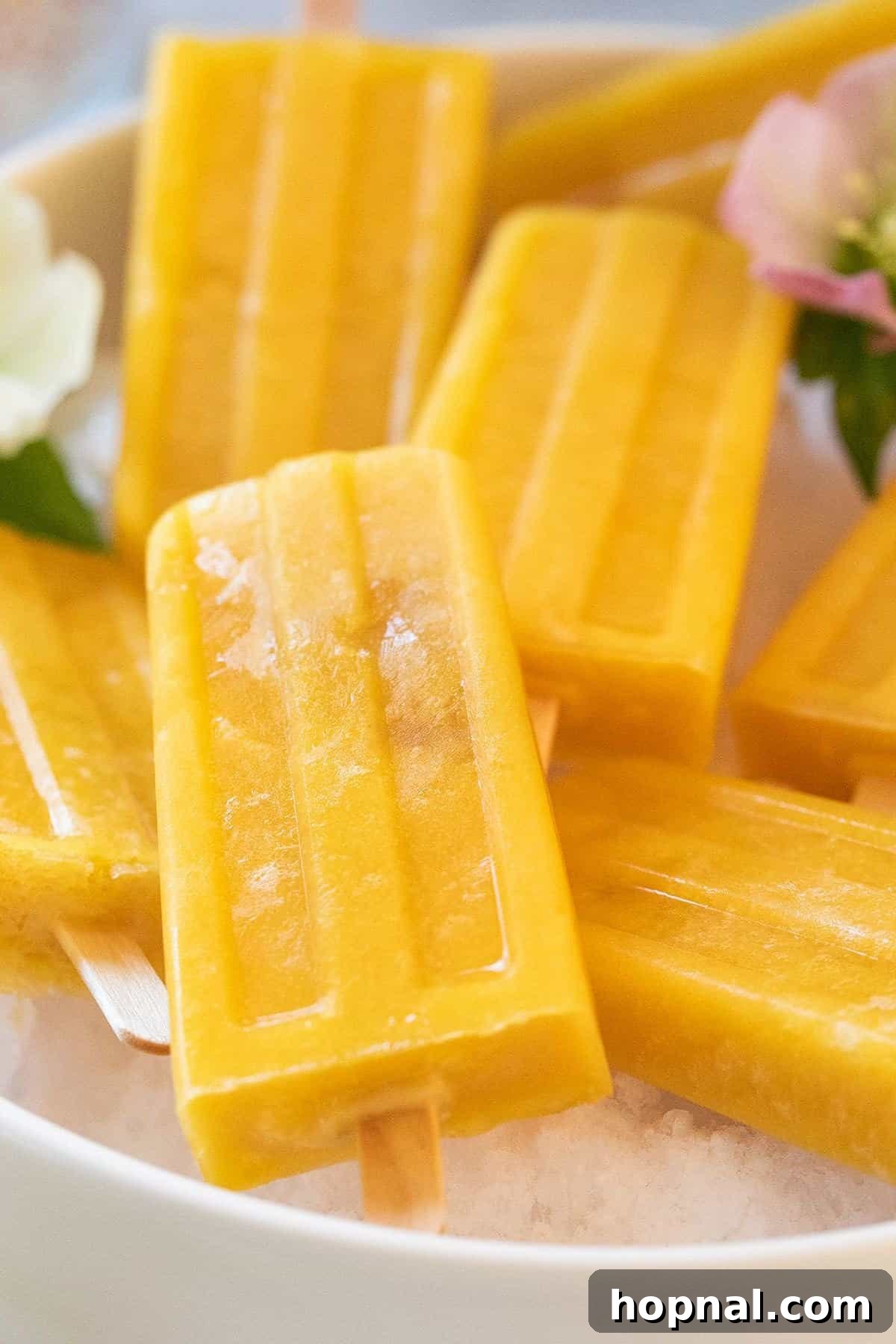 A bright yellow homemade mango popsicle held against a blurred background of fresh mango slices, highlighting its vibrant color and tropical essence.