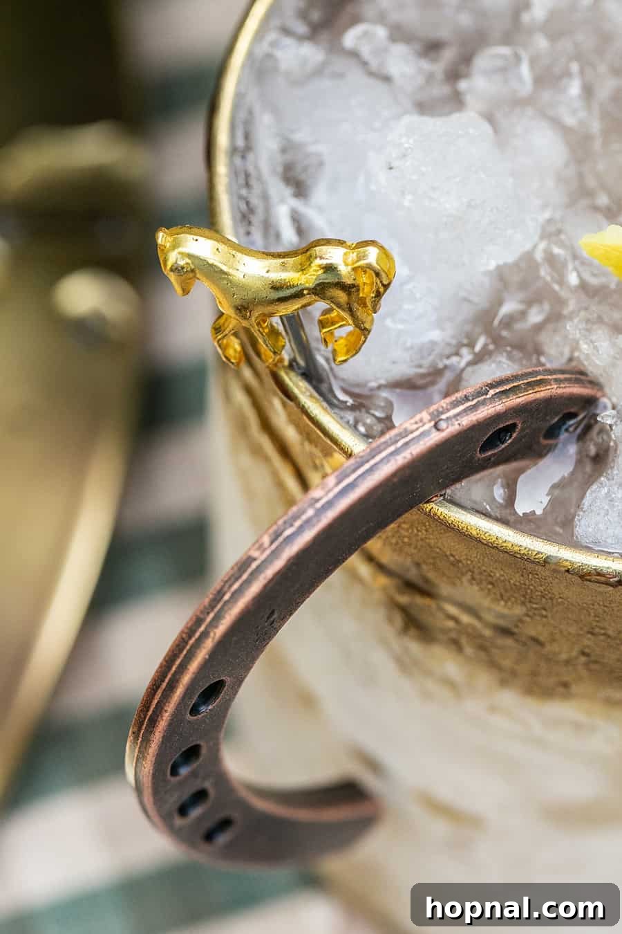 A horse-shaped cocktail pick garnishing a cocktail in a gold hammered mug, a charming Derby detail.
