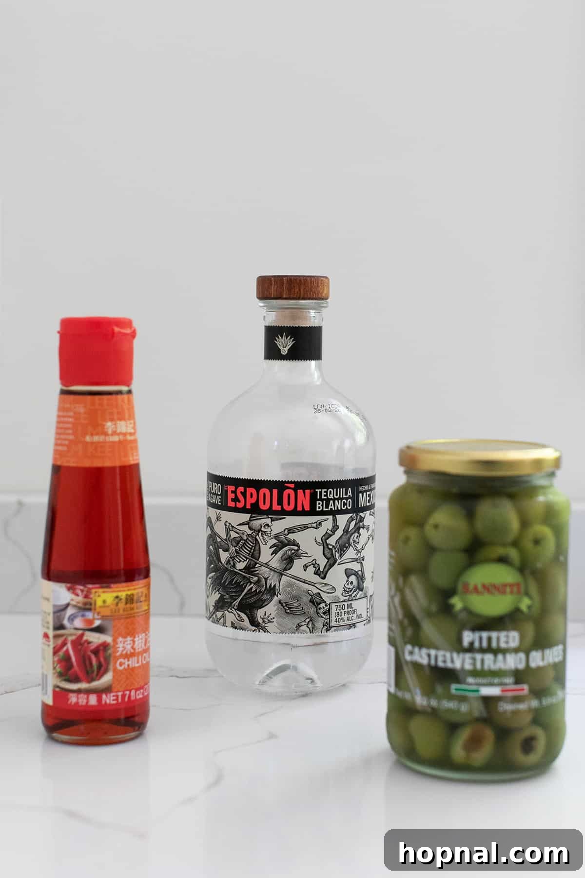 Tequila's Dirty Martini 3 The three essential ingredients for a Tequila Dirty Martini are displayed: a bottle of premium Espolòn Blanco tequila, a bowl brimming with bright green olives, and a small dish containing vibrant red chili oil, all artfully arranged on a clean white surface.