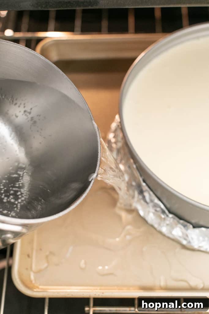 Irresistible Homemade Cheesecake 8 A cheesecake in a foil-wrapped springform pan, placed within a larger roasting pan, with hot water being poured to create a crucial water bath for even baking.