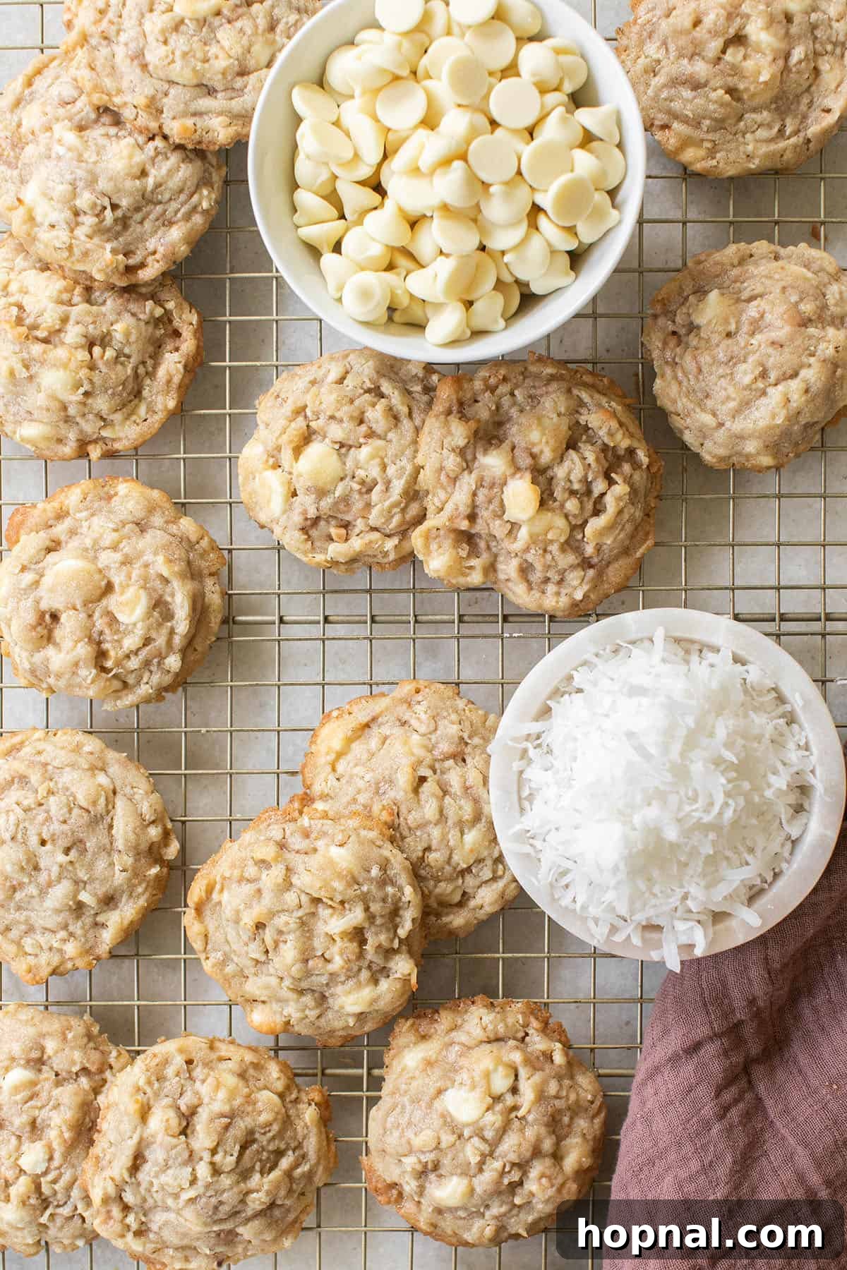 Ultimate Kitchen Sink Cookies 6 Freshly baked Kitchen Sink Cookies cooling on a wire rack, with bowls of white chocolate chips and shredded coconut in the background, hinting at their delicious contents.